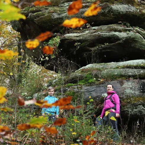 Eine beeindruckende Felsformation umgeben von bunten Herbstblättern. Zwei Personen stehen in der Natur und genießen die Landschaft.
