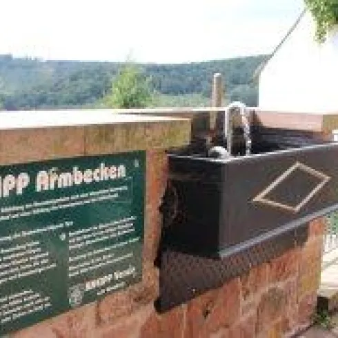 Ein Kneipp-Armbad mit einem Wasserhahn und einer erklärenden Tafel. Im Hintergrund ist eine grüne Landschaft zu sehen.