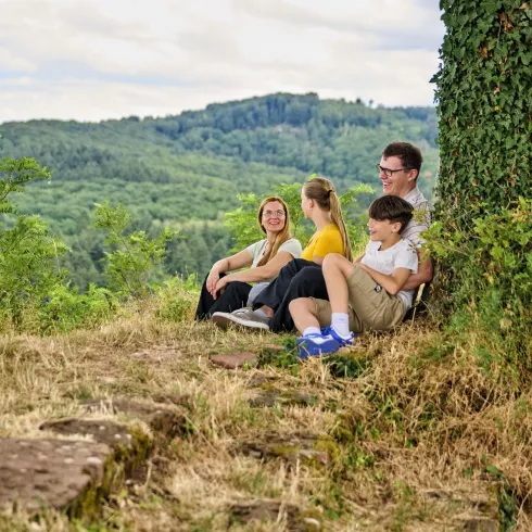 Eine Gruppe von vier Personen sitzt entspannt auf dem Boden in der Natur. Im Hintergrund sind sanfte Hügel und Bäume sichtbar.