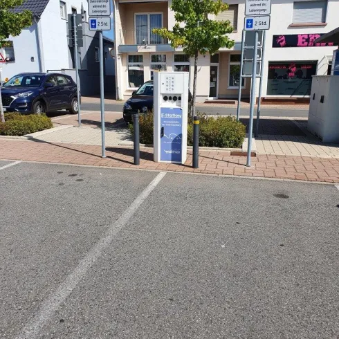 Ein Parkplatz mit Parkautomaten und angrenzenden Parkplätzen. Im Hintergrund sind einige Gebäude und Pflanzen sichtbar.