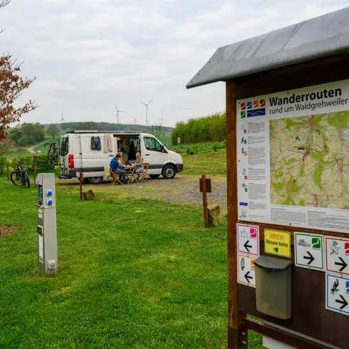 Eine Wiese mit einem Campingvan und einem Wegweiser zu Wanderwegen. Im Hintergrund sind einige Bäume und sanfte Hügel zu sehen.
