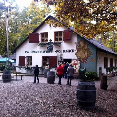 Ein gemütliches Restaurant im Wald mit einer einladenden Terrasse. Einige Gäste stehen vor dem Gebäude und genießen die natürliche Umgebung.