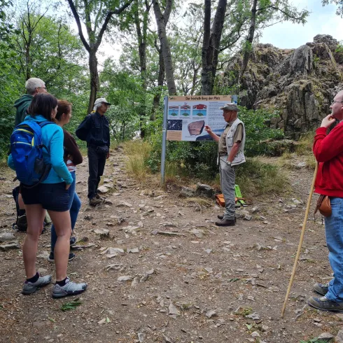 Eine Gruppe von Wanderern hört einem Tourführer zu, der an einem Informationsschild steht. Im Hintergrund sind Bäume und Felsen zu sehen.