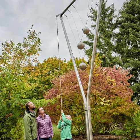 Eine Gruppe von drei Personen steht an einem modernen Spielgerät im Freien. Im Hintergrund sind bunte Bäume sichtbar, die eine herbstliche Atmosphäre schaffen.
