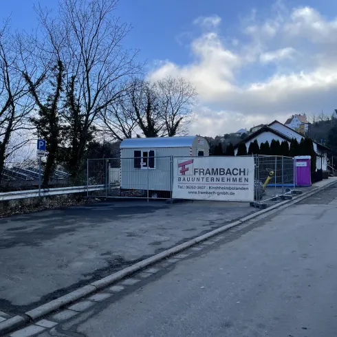 Ein Baustellenbereich mit einem Wohncontainer und einem Werbebanner. Im Hintergrund sind einige Bäume und Wohnhäuser zu sehen.