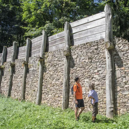 Eine alte Mauer aus Stein mit Holzpfosten in einer grünen Umgebung. Zwei Personen betrachten die Struktur in der Natur.