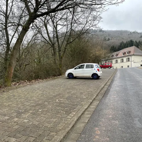 Ein kleiner, weißer Wagen steht an einer unbefahrenen Straße. Im Hintergrund sind Bäume und ein Gebäude zu sehen.