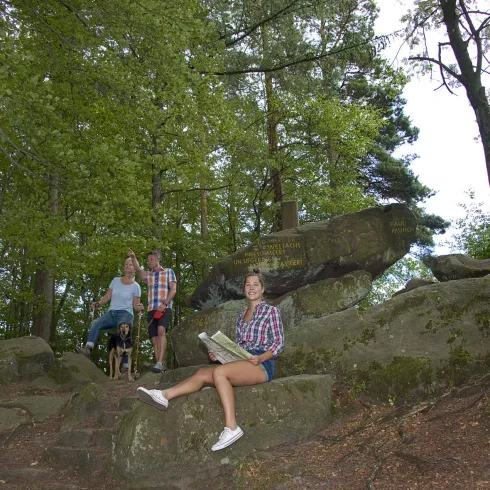 Eine Gruppe von Menschen steht in einem Wald, umgeben von Bäumen und Felsen. Eine Person sitzt auf einem Stein und hält ein Buch in der Hand.