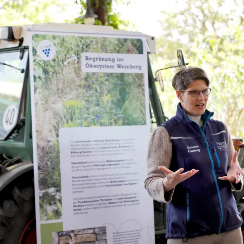 Eine Frau spricht neben einem Traktor und einem Informationsplakat. Das Plakat thematisiert die Begrünung im Ökosystem Weinberg.