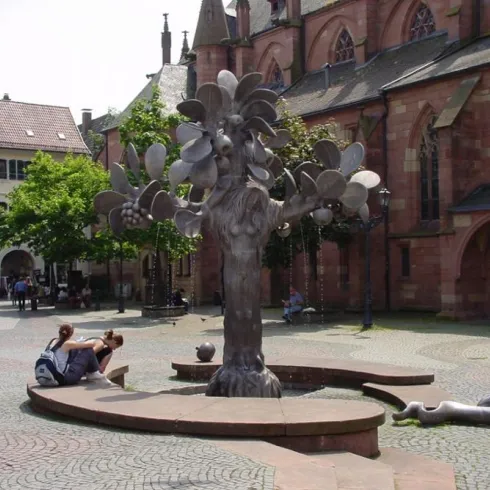 Ein kunstvoller Baum aus Metall steht im Mittelpunkt eines Platzes, umgeben von Bänken und Bäumen. Im Hintergrund sind historische Gebäude und Passanten zu sehen.