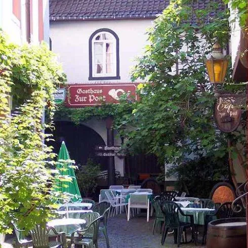 Ein malerischer Innenhof mit Tischen und Stühlen, umgeben von grünen Pflanzen. Im Hintergrund ist ein Gasthaus mit einem einladenden Schild zu sehen.