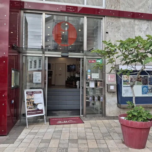 Ein modernes Gebäude mit einer gläsernen Eingangstür. Vor der Tür steht ein roter Blumentopf mit einer Pflanze.