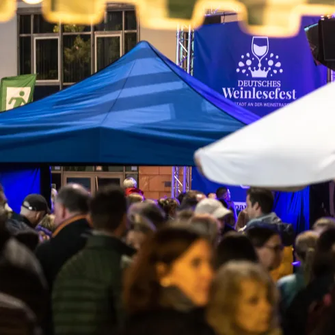 Ein lebendiges Weinfest mit vielen Menschen und einem blauen Zelt. Im Hintergrund ist ein Banner mit dem Schriftzug „Deutsches Weinfest“ sichtbar.