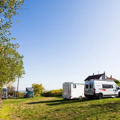 Ein ruhiger Campingplatz mit Wohnmobilen und einem kleinen Haus. Umgeben von Bäumen und einer klaren blauen Himmel.