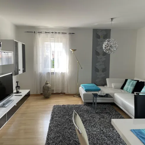 Ein modernes Wohnzimmer mit einem weißen Sofa und bunten Kissen. Der Raum ist hell und freundlich mit einem großen Fenster und einer stilvollen Dekoration.