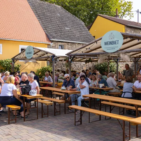 Eine lebhafte Außensitzfläche mit vielen Gästen, die an langen Tischen sitzen. Im Hintergrund sind bunte Gebäude und grüne Pflanzen zu sehen.