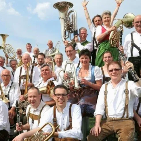Eine Musikkapelle mit vielen Mitgliedern in traditioneller Tracht. Sie posieren fröhlich im Freien mit verschiedenen Instrumenten.