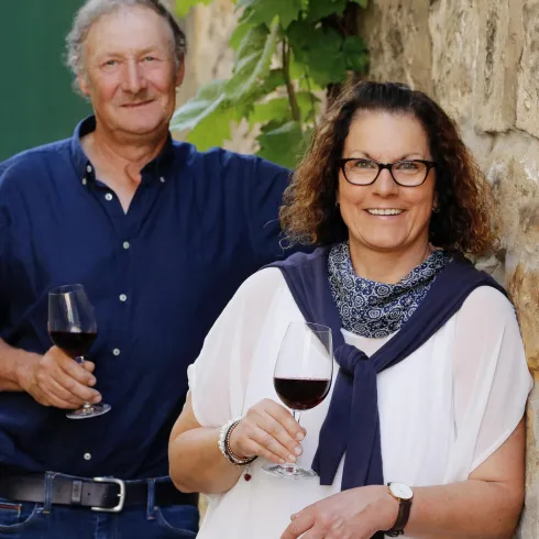 Ein Paar steht lächelnd vor einer Steinmauer. Beide halten ein Glas Rotwein in der Hand.