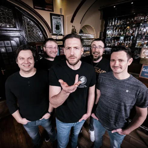 Eine fünfköpfige Band steht lächelnd in einer Bar vor einer Theke mit Getränken. Die Atmosphäre ist entspannt und freundlich.