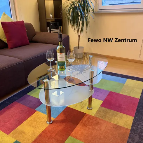 Ein stilvolles Wohnzimmer mit einem modernen, gläsernen Couchtisch, einer Flasche Wein und zwei Gläsern. Bunte Teppichmuster und gemütliche Sofakissen sorgen für eine einladende Atmosphäre.
