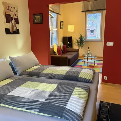 Ein gemütliches Zimmer mit einem Doppelbett und bunten Kissen. Im Hintergrund befindet sich ein Wohnzimmerbereich mit einer Couch und einem bunten Teppich.