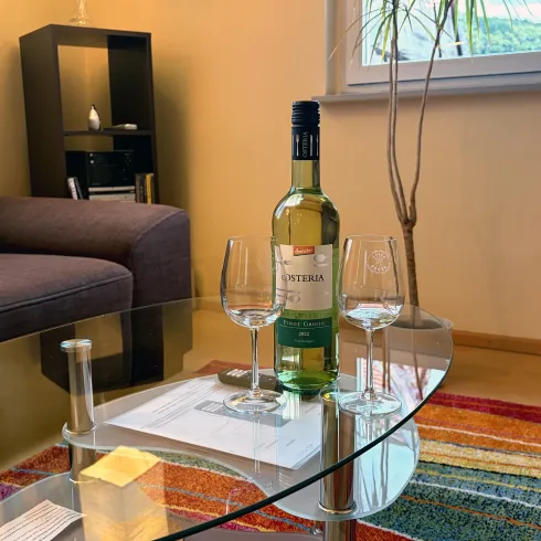Ein gemütlicher Wohnraum mit einem Glas-Esstisch. Auf dem Tisch stehen eine Flasche Wein und drei Glasbecher.