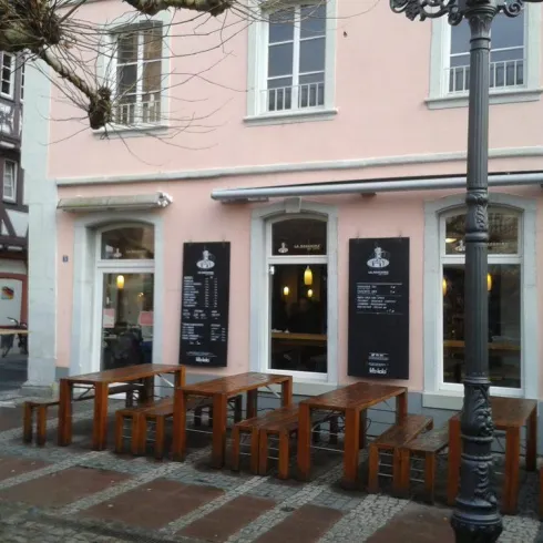 Ein gemütliches Café mit Holztischen und -bänken im Freien. Die Fassade ist in einem hellen Rosa gestrichen und hat große Fenster.
