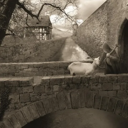 Eine sepiafarbene Landschaft mit einer alten Steinbrücke und einem Hirten, der ein Schaf streichelt. Im Hintergrund sind ein historisches Haus und eine Mauer zu sehen.