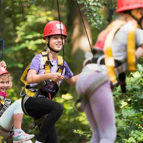 Eine Gruppe von Kindern schwingt fröhlich an Seilen im Wald. Sie tragen Helme und Sicherheitsgeschirr.