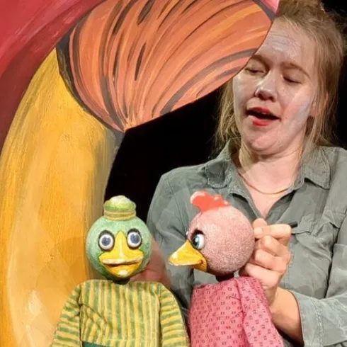 Eine Frau mit geschminktem Gesicht spielt mit zwei Handpuppen. Im Hintergrund ist ein großes, buntes Pilz-Motiv zu sehen.