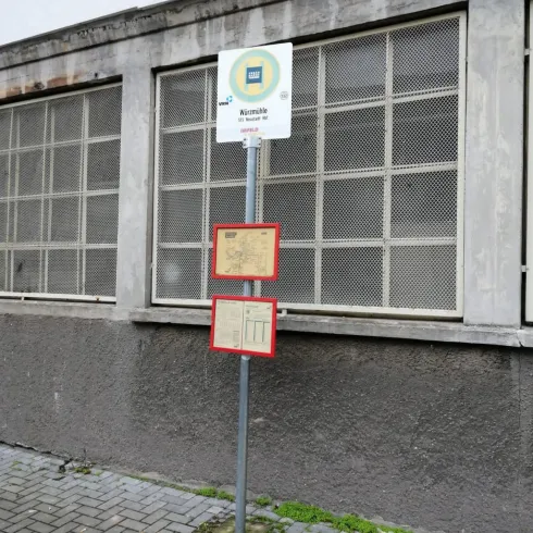 Eine Haltestelle mit einem Schild und Fahrplänen. Die Wand hinter der Haltestelle ist grau und die Fenster sind mit Gitter versehen.