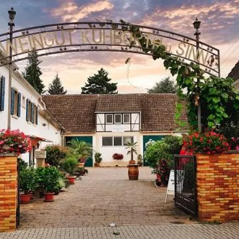 Ein malerischer Weingut-Eingang mit blühenden Pflanzen und einem einladenden Torbogen. Im Hintergrund sind charmante Gebäude und ein sanfter Himmel zu sehen.