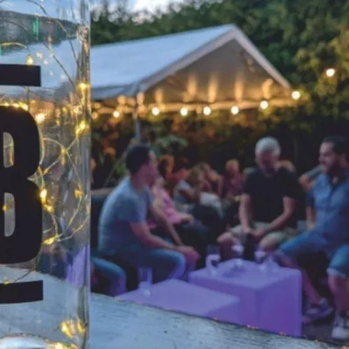 Ein Glas mit Lichtern steht im Vordergrund, während im Hintergrund Menschen in einer geselligen Runde sitzen. Die Atmosphäre ist festlich und entspannt.