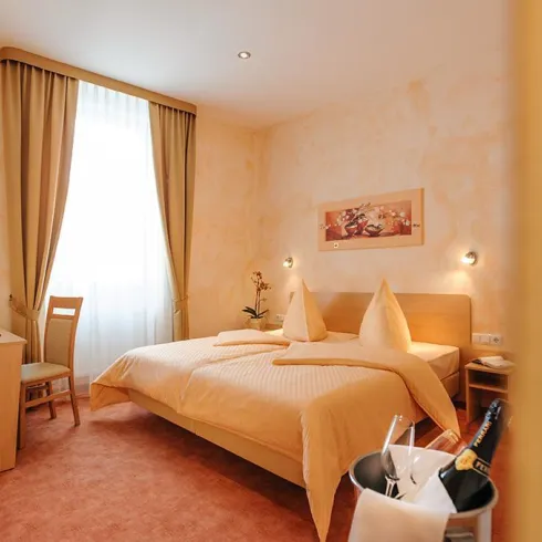 Ein gemütliches Hotelzimmer mit einem Doppelbett und einem kleinen Tisch. Die Wände sind hell, und das Zimmer hat große Fenster mit Vorhängen.