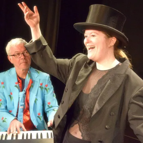 Eine fröhliche Aufführung mit einer Sängerin in einem eleganten Outfit und Zylinder. Im Hintergrund spielt ein Musiker am Klavier.