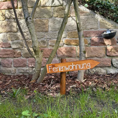 Ein Holzschild mit der Aufschrift "Ferienwohnung" zeigt in Richtung des Weges. Im Hintergrund ist eine Steinmauer und ein kleiner Garten sichtbar.