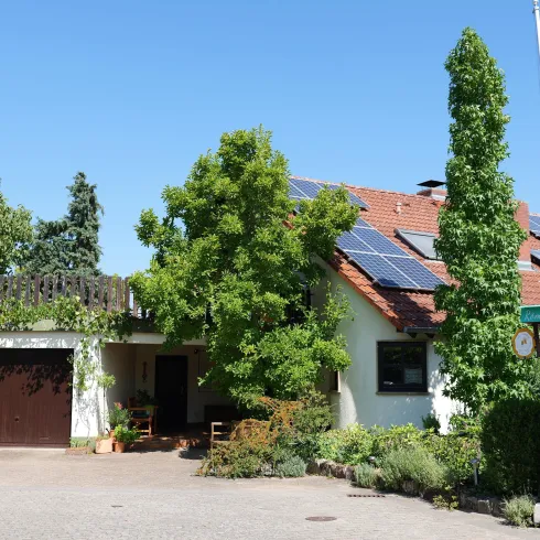 Ein Einfamilienhaus mit einem roten Dach und Solarpanelen. Es ist von grünen Bäumen und Pflanzen umgeben und steht unter einem klaren blauen Himmel.
