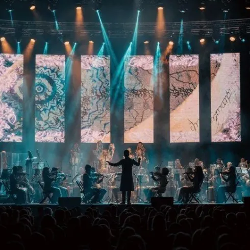 Ein Orchester spielt auf einer Bühne, während ein Dirigent das Ensemble leitet. Im Hintergrund sind große Leinwände mit bunten Mustern und Lichtern zu sehen.