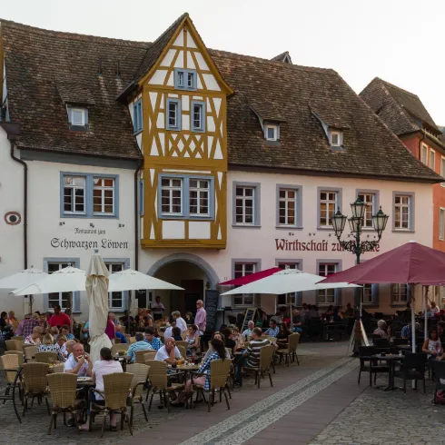 Eine malerische Straße mit traditioneller Architektur und einem Restaurant im Freien. Viele Menschen genießen ihre Zeit am Tisch unter Sonnenschirmen.