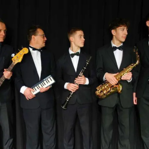 Eine Gruppe von fünf Musikern steht nebeneinander. Sie tragen elegante Anzüge und halten verschiedene Musikinstrumente.