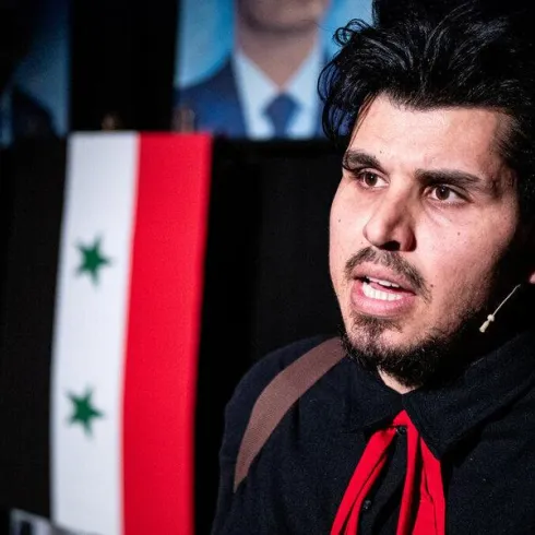 Ein Mann spricht passionately, während er vor einer syrischen Flagge steht. Im Hintergrund sind Bilder von Männern sichtbar.