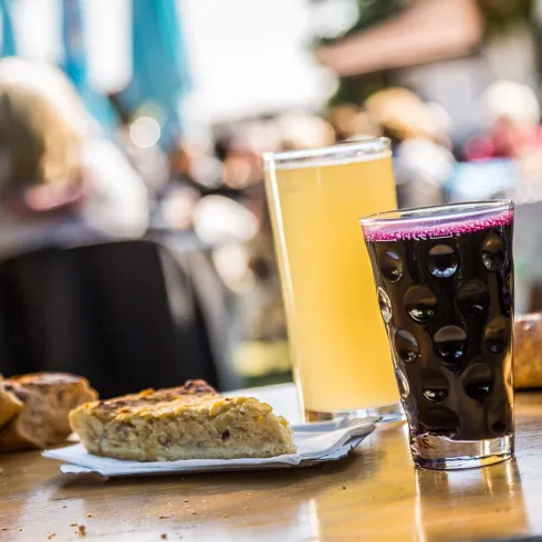 Ein Tisch mit zwei Getränken, einem Bier und einer dunklen Limonade, sowie verschiedenen Backwaren. Im Hintergrund sind unscharfe Personen und eine sonnige Atmosphäre zu sehen.