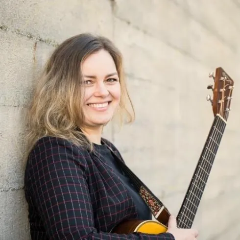 Eine Frau mit langen, welligen Haaren lächelt und steht an einer Wand. Sie hält eine Gitarre und trägt ein schwarzes, kariertes Outfit.