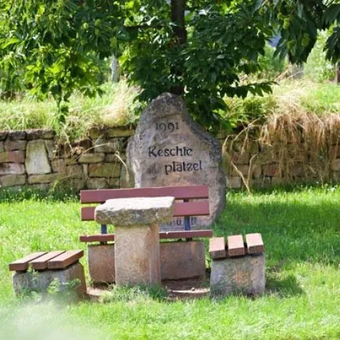 Ein gemütlicher Platz im Freien mit einem Tisch und Bänken aus Stein und Holz. Im Hintergrund steht ein große Stein mit der Aufschrift "Keschte Platzel".