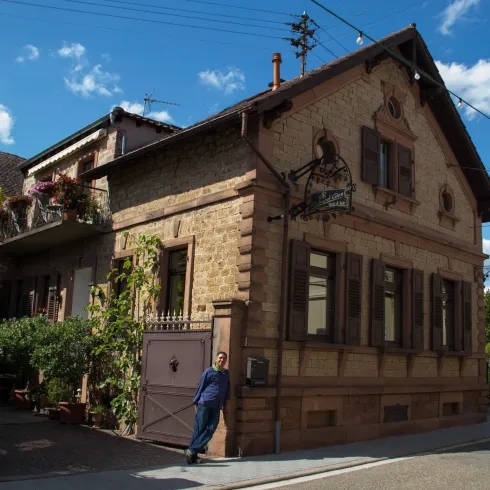 Ein schönes, historisches Gebäude aus Stein mit Fenstern in Holzrahmen. Vor dem Haus steht eine Person, umgeben von grünen Pflanzen.
