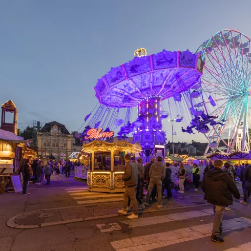 Ein festlicher Jahrmarkt mit bunten Fahrgeschäften und vielen Menschen. Im Hintergrund sind das Riesenrad und die beleuchtete Karusselle zu sehen.