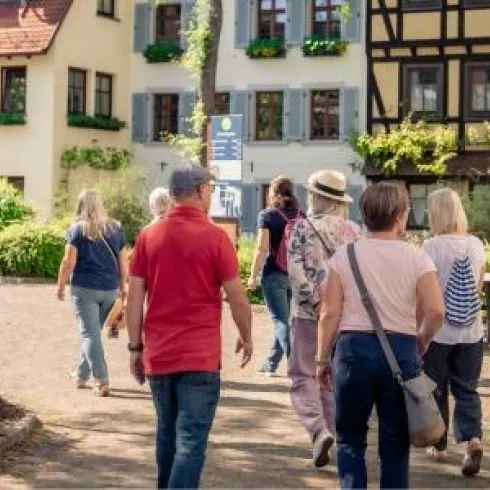 Eine Gruppe von Menschen spaziert durch eine malerische Straße mit traditionellen Häusern. Es ist sonnig und die Umgebung ist grün und einladend.
