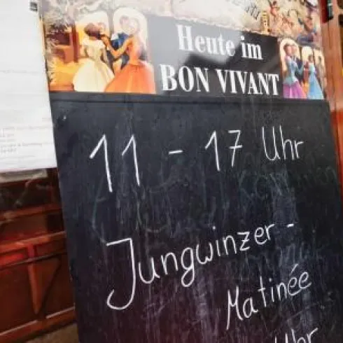 Ein schwarzes Brett mit Informationen über die Öffnungszeiten und eine Veranstaltung. Es steht "Heute im BON VIVANT" und "Jungwinzer - Matinée" geschrieben.
