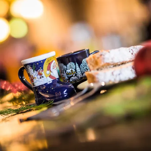 Zwei Tassen stehen auf einem Tisch neben leicht geschnittenem Brot. Im Hintergrund sind unscharfe Lichter zu sehen, die eine gemütliche Atmosphäre schaffen.