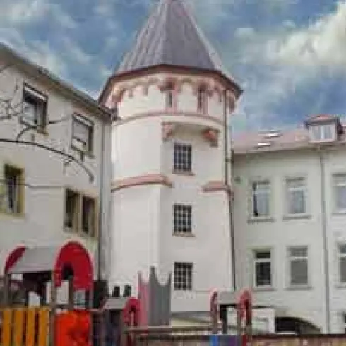 Ein historischer Turm mit einem spitzen Dach, umgeben von modernen Gebäuden. Im Vordergrund befindet sich ein Spielplatz mit bunten Spielgeräten.